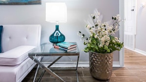 Una sala de estar moderna con un sofá blanco, una mesa de centro de cristal con libros, una lámpara color turquesa y una planta en maceta.