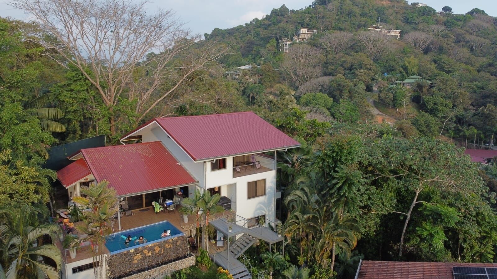 A two-story house with a red roof, a swimming pool, and a view of a lush, green hillside.