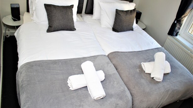 A neatly made bed with white and gray linens, two pillows, and two rolled towels on gray bed runners. - CITY CENTRE SPACIOUS 5 BEDROOMS HOUSE AYLESBURY (Buckinghamshire)