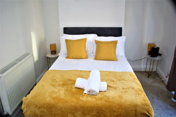 A hotel room with a bed, two bedside tables, a radiator, and a folded towel on the bed.