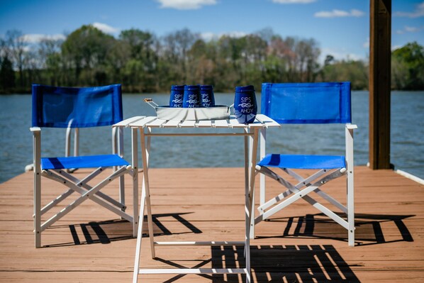 Una terraza de madera con una mesa y dos sillas plegables azules, un toldo azul y cuatro contenedores azules con la etiqueta 'SIPS AHOY' frente a un lago.