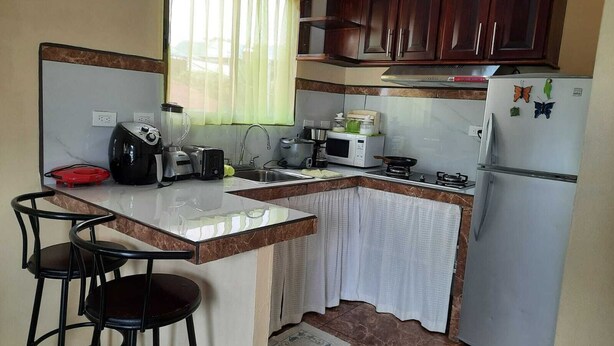 Una cucina con un piano di lavoro in marmo, un lavello, un fornello, un forno a microonde, un frigorifero e una finestra con tende. Sul piano di lavoro ci sono tre sgabelli neri e un elettrodomestico rosso.