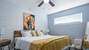 Un dormitorio con cama, mesita de noche, ventilador de techo, ventana con persianas y cesta de ropa.