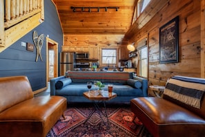 Interior acogedor de una cabaña con techo de madera, una zona de cocina y un área de estar con un sofá azul y otomanas de cuero.