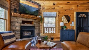 A cozy cabin interior with a stone fireplace, leather sofas, a small table with drinks, and a TV mounted on the wall.