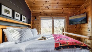 A wooden cabin bedroom with a bed, framed pictures, a TV, and a window with a view.