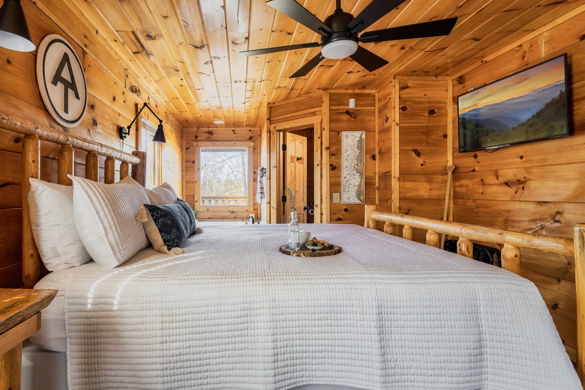 Un dormitorio de cabaña de madera con una cama, un ventilador de techo, un televisor y un ventanal grande.