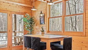 L’intérieur d’un chalet en bois comprend une table à manger, quatre chaises, une plante en pot et de grandes fenêtres donnant sur la forêt.