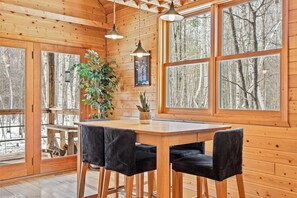 A wooden cabin interior with a dining table, four chairs, a potted plant, and large windows offering a view of the forest.