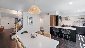 A modern kitchen with a central island, dark wooden flooring, and a dining area with a white table and wicker chairs. - ★Golf & Couples Oasis★ Midcentury w/6 Pools, Hot Tub (Palm Springs)