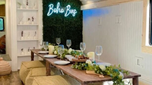 A dining area with a long wooden table set for a meal, surrounded by cushioned benches, and a neon sign reading "Boho Bus" on the wall. - Gentry Pines: Huge backyard ~ Loaded Game Room (Vestavia Hills)