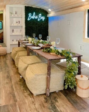 Un comedor con una larga mesa de madera dispuesta para una comida, rodeada de bancos acolchados, y un letrero de neón en la pared que dice "Boho Bus".