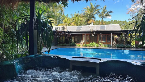 A swimming pool with a hot tub, surrounded by lush tropical plants and palm trees, with a modern house in the background.
