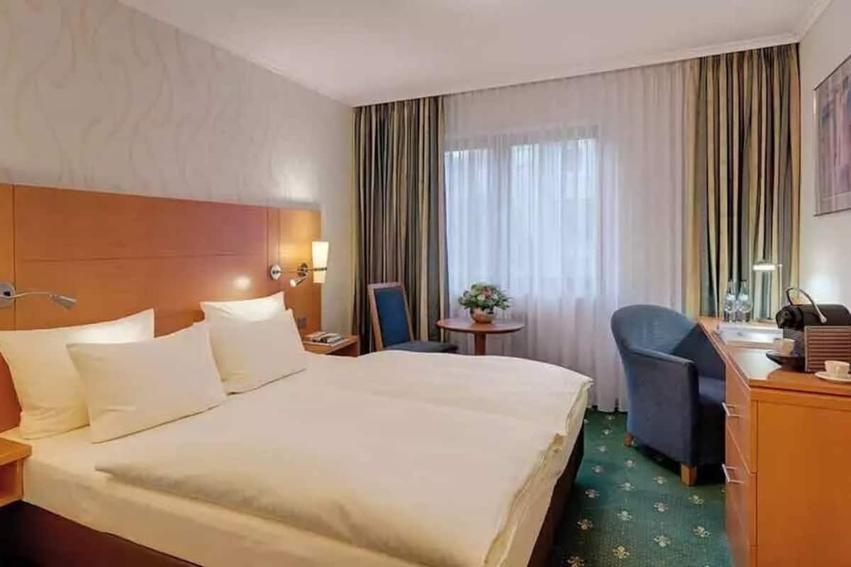 Une chambre d’hôtel avec un grand lit, un bureau avec une chaise, une petite table avec une plante et des rideaux à la fenêtre.