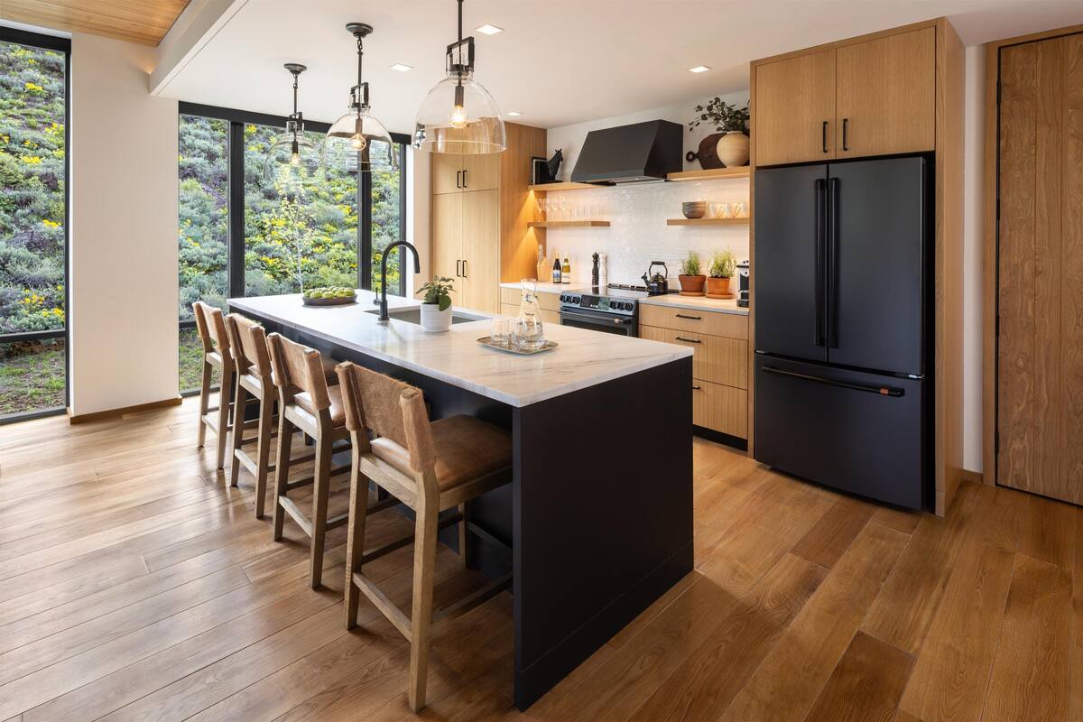 A modern kitchen with a large island, wooden chairs, a black refrigerator, and a view of greenery outside.