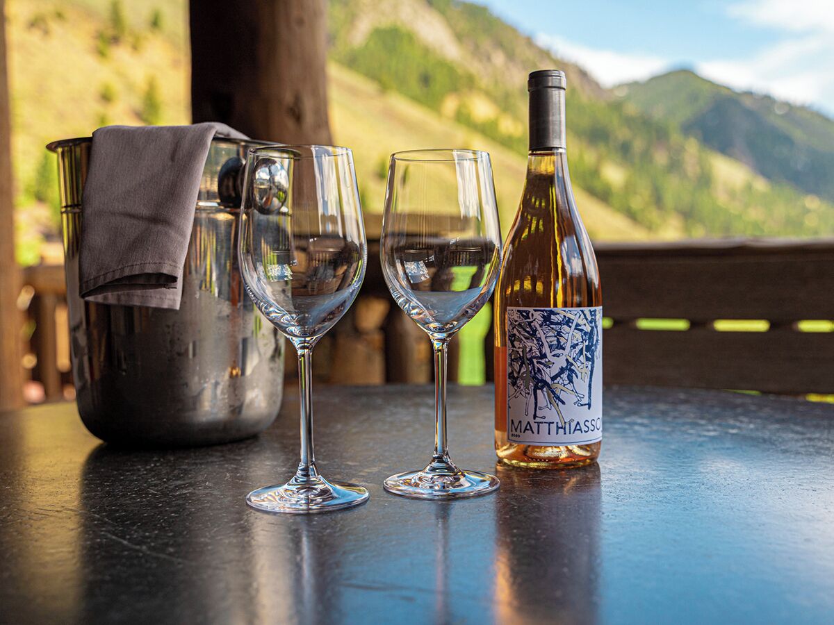A bottle of Matt Hiasch wine, two glasses, and a metal bucket on a table with a mountain view in the background.