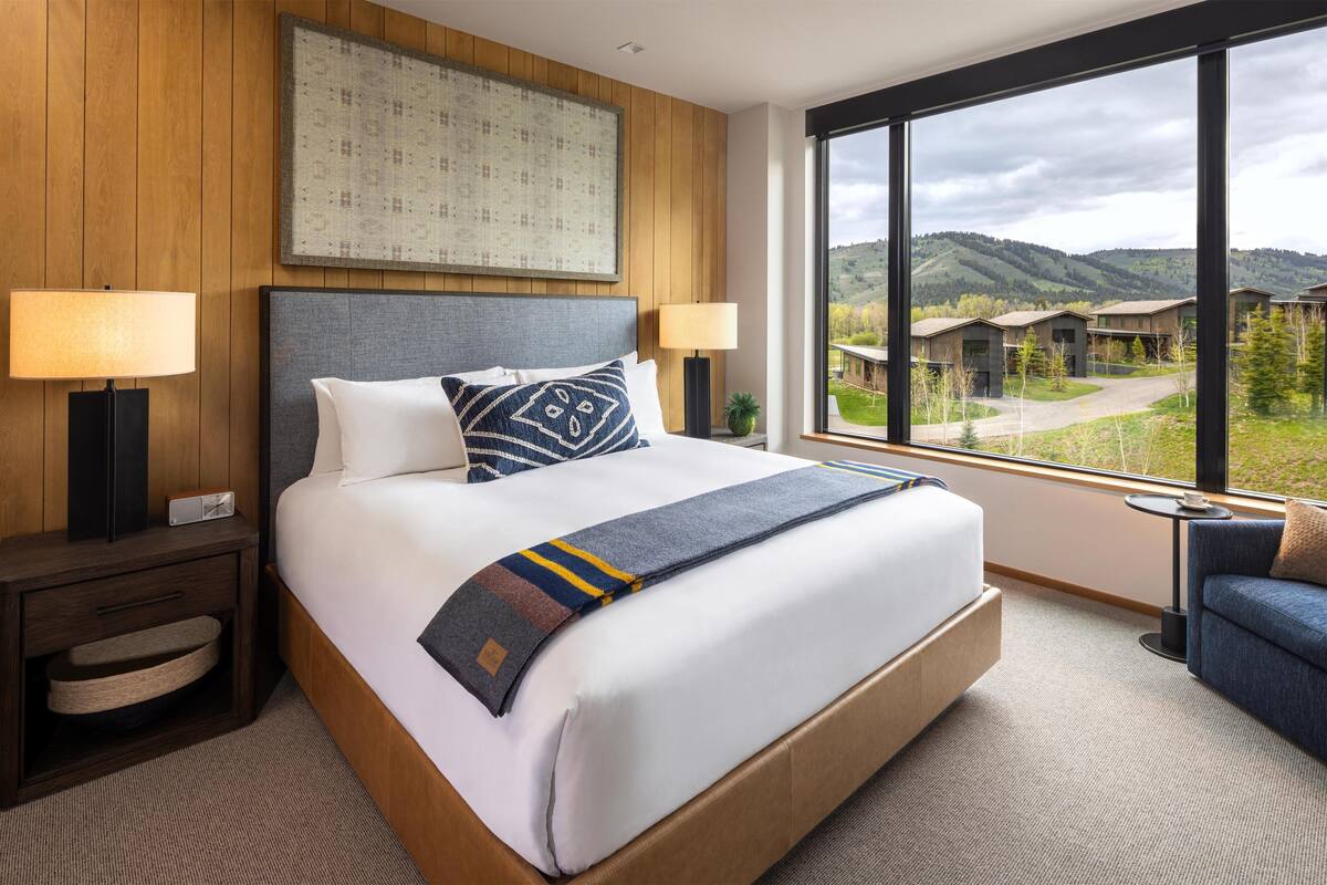 A hotel room with a large bed, two bedside lamps, a nightstand, a sofa, and a window with a view of the hotel and surrounding hills.