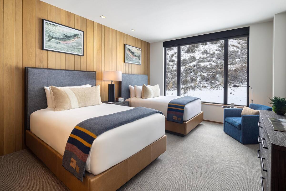 A modern hotel room with two beds, a desk, and large windows offering a view of a snowy landscape.