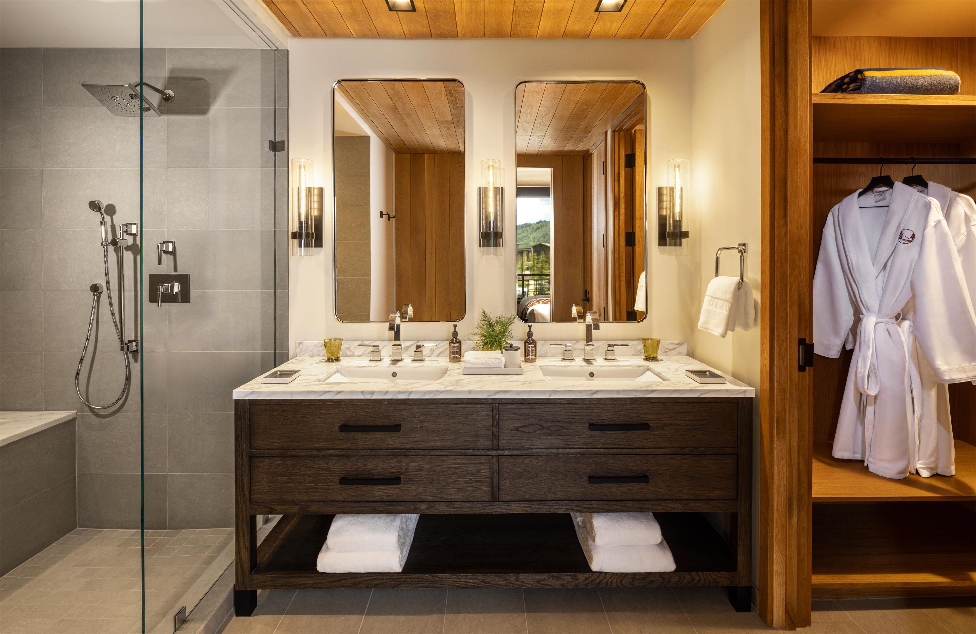 Une salle de bain moderne dotée d’une douche à parois de verre, d’un meuble-lavabo en bois avec deux vasques et d’un dressing où sont rangés des peignoirs.