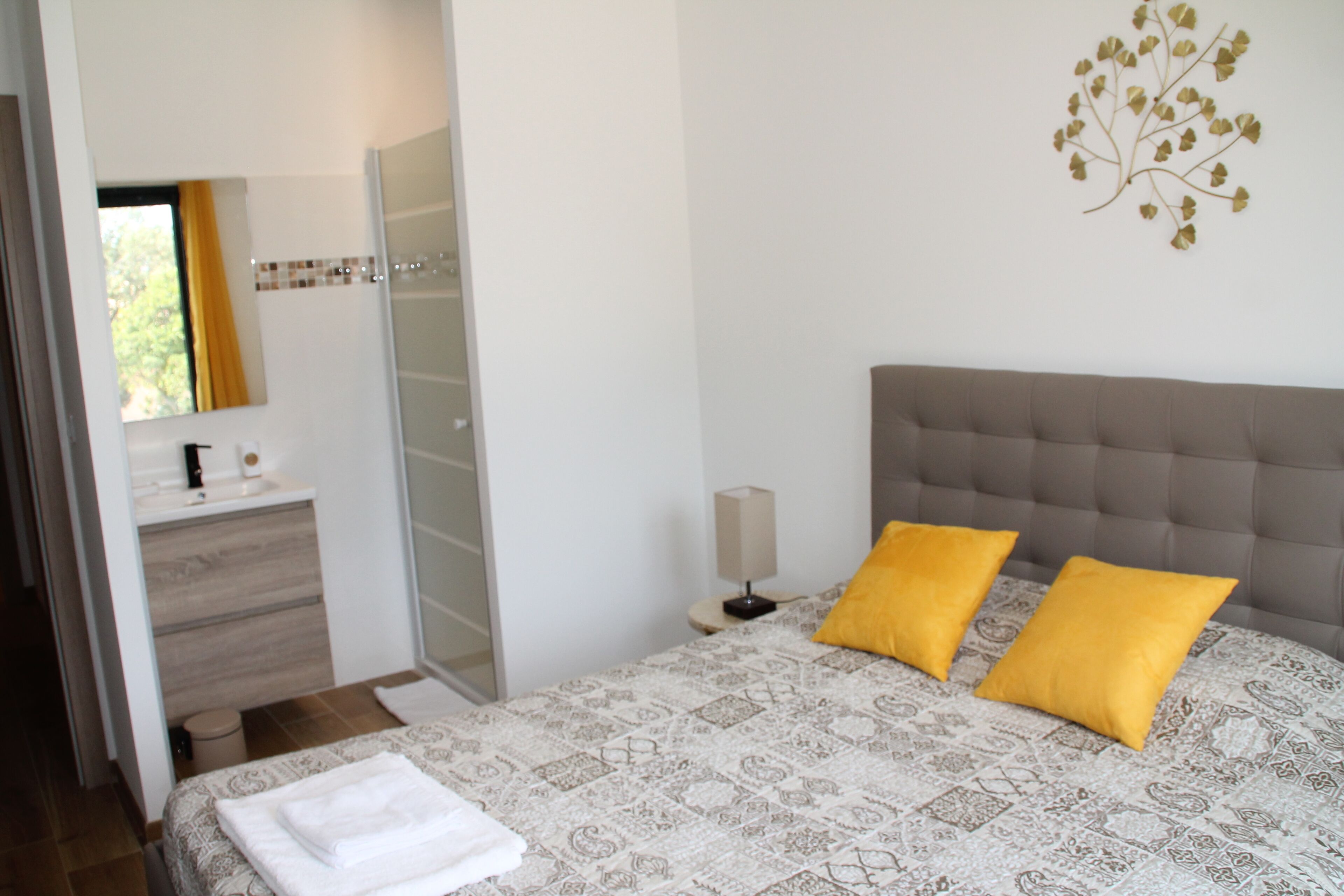 A bedroom with a bed, two pillows, a bedside table, a lamp, a mirror, a window, and a bathroom with a sink and a towel.