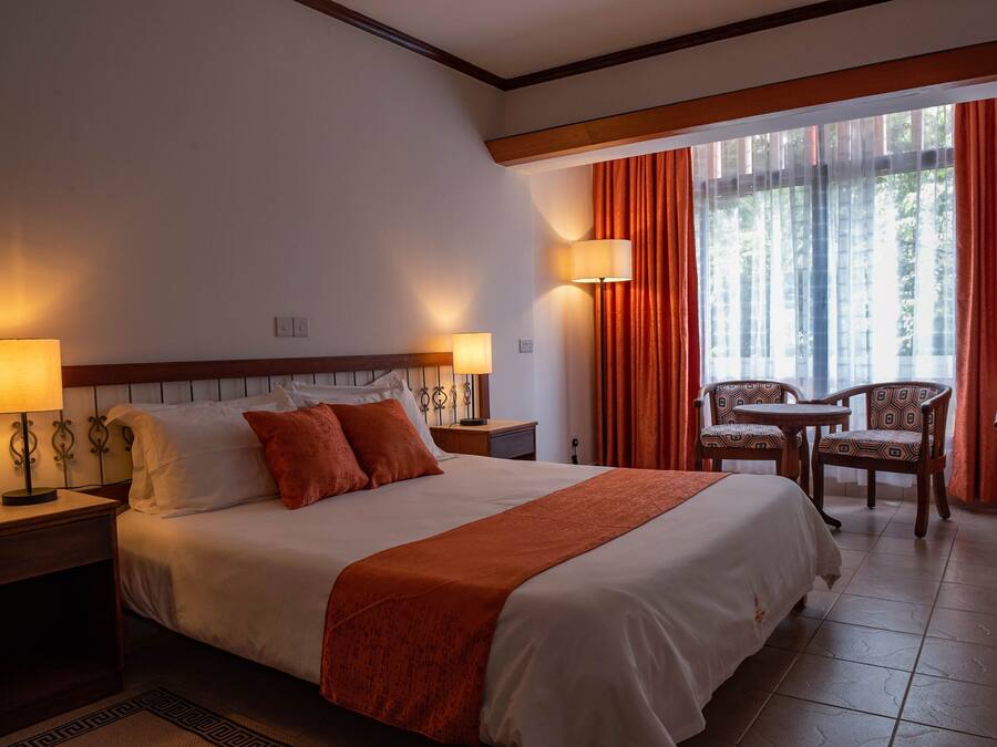 Une chambre d’hôtel avec un grand lit, deux lampes de chevet, une petite table avec des chaises et une fenêtre avec des rideaux.