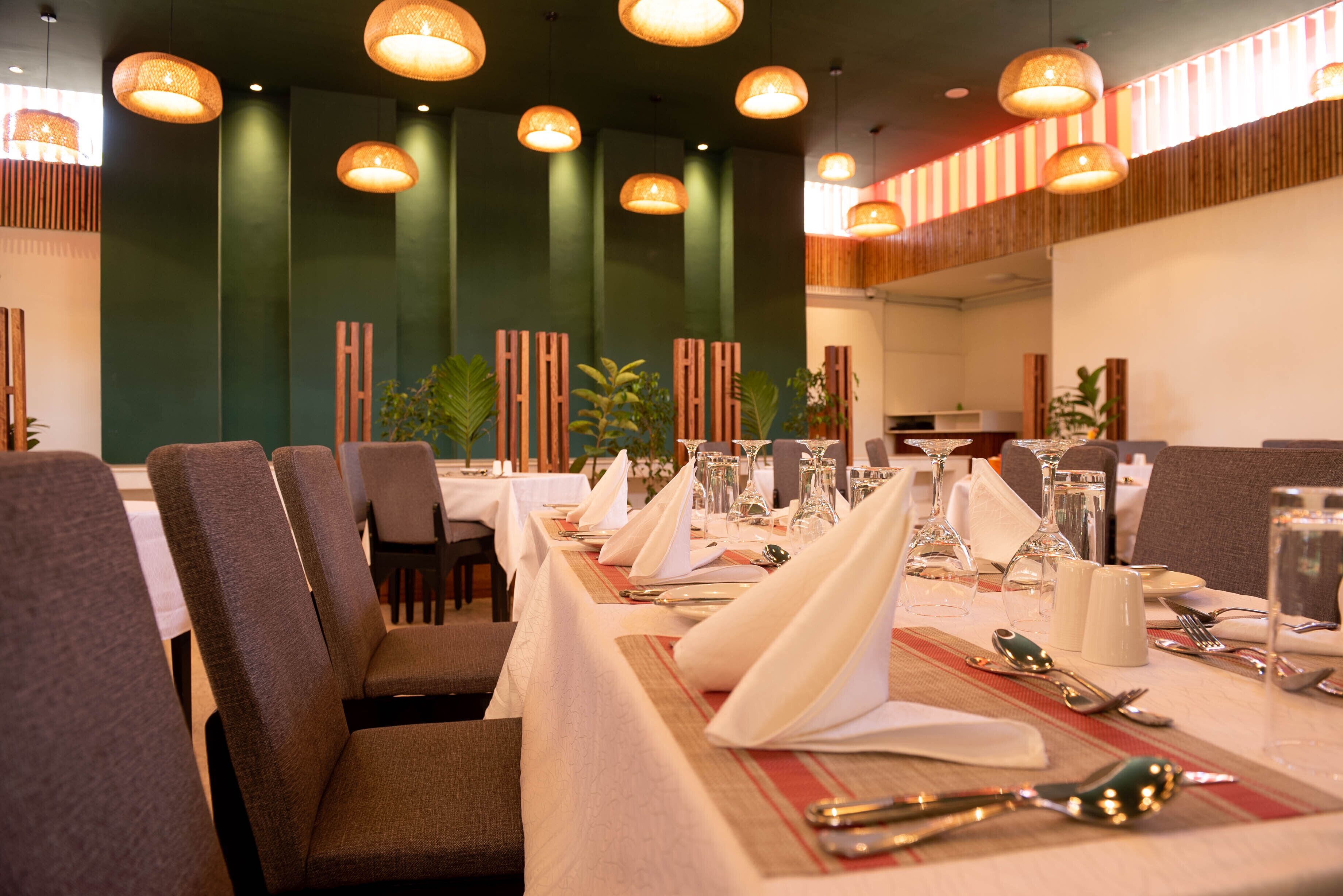 A restaurant with long tables set for dining, featuring a modern interior with green walls and hanging pendant lights.