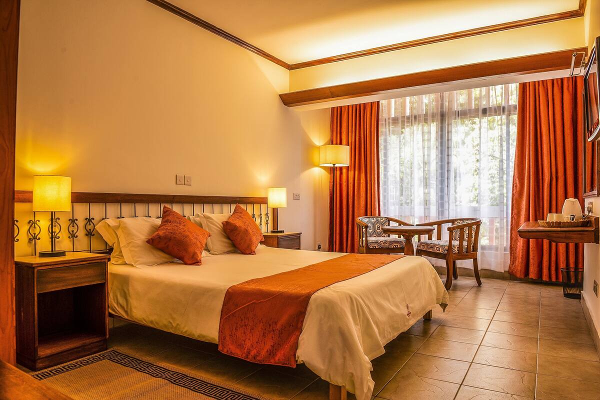 Une chambre d’hôtel avec un grand lit, deux tables de chevet avec des lampes, une chaise, une petite table et une fenêtre avec des rideaux.