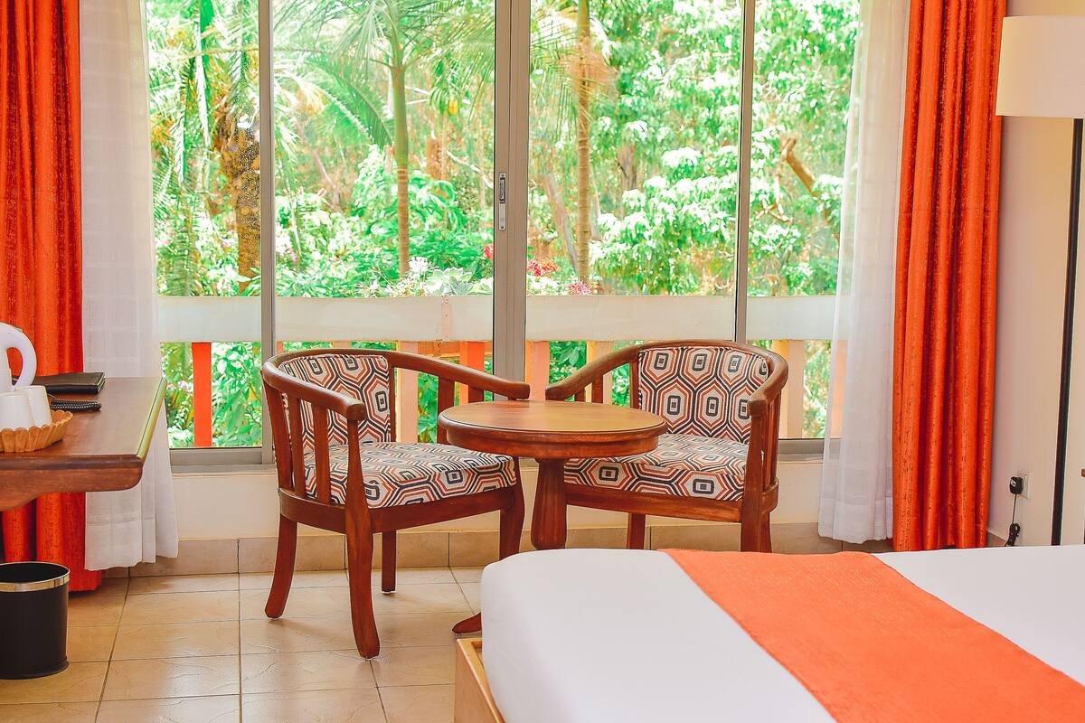 Une chambre avec un lit, une table entourée de chaises et une vue sur une végétation luxuriante, visible à travers de grandes fenêtres.