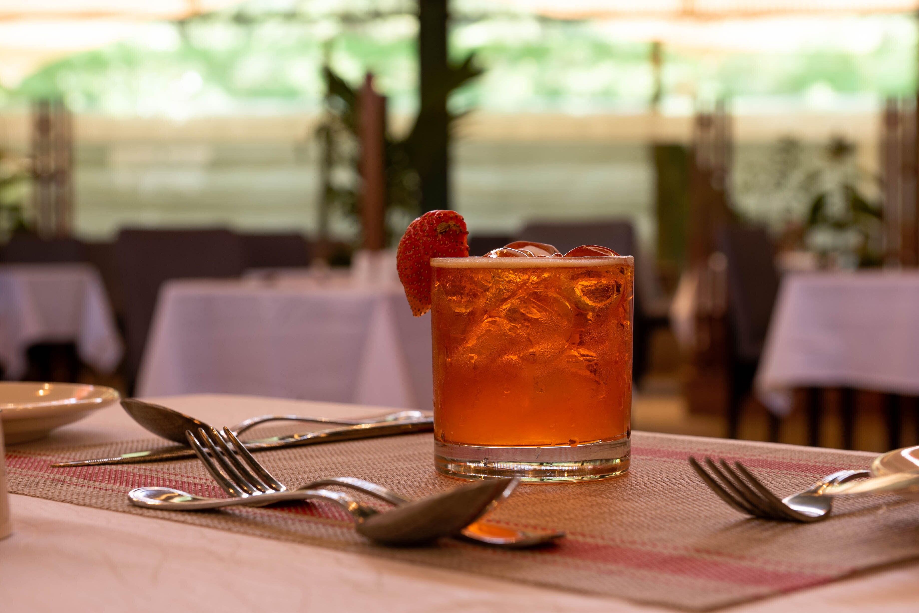 A glass of amber-colored beverage with a strawberry garnish on a table with cutlery.