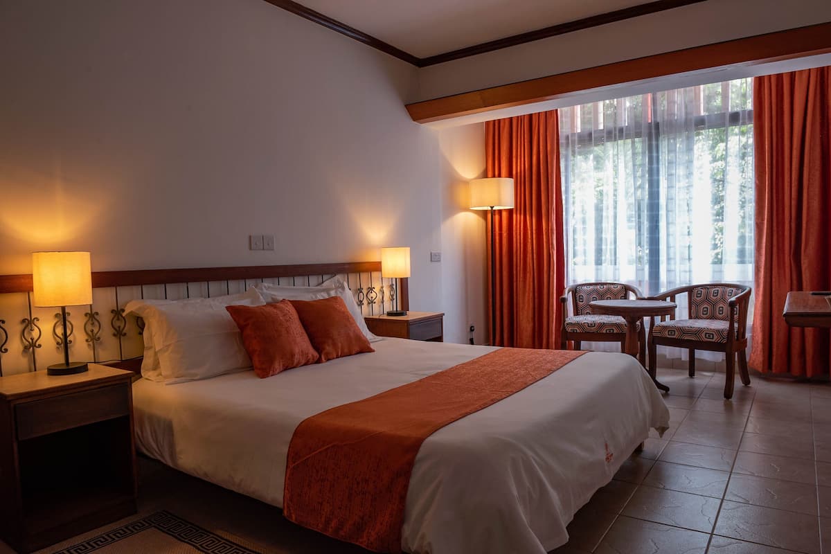 Une chambre d’hôtel avec un grand lit, deux lampes de chevet, une petite table avec des chaises et une vue sur l’extérieur à travers les rideaux.