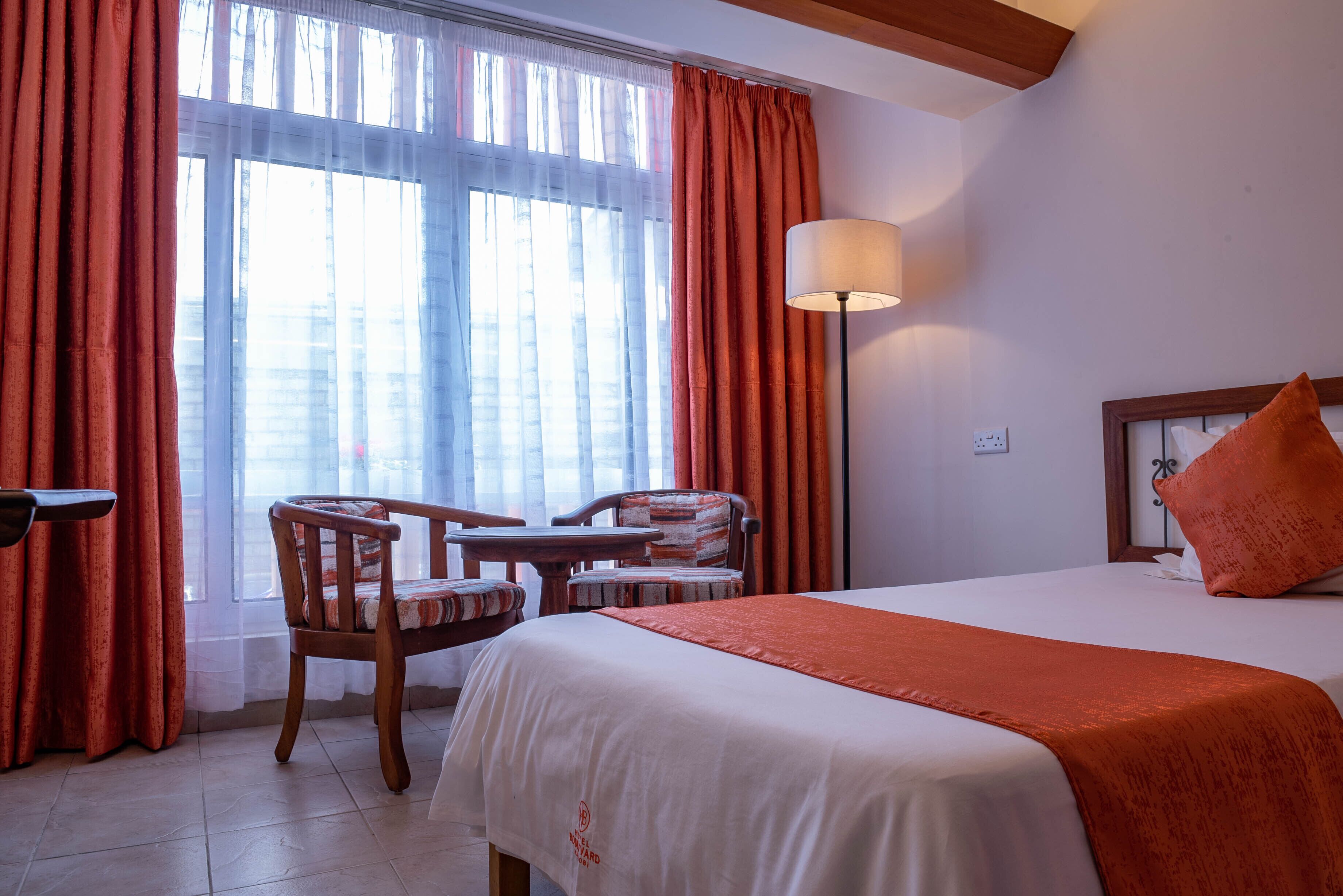 A hotel room with a bed, a desk, two chairs, a lamp, and a window with curtains.
