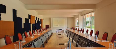 Un salón de conferencias con una larga mesa, sillas rojas, botellas de agua y una pantalla de proyector que muestra