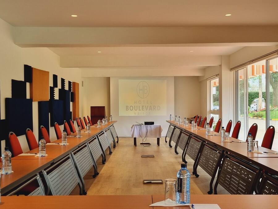 A conference room with a long table, red chairs, water bottles, and a projector screen displaying "HOTEL BOULEVARD".
