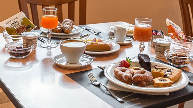 Una mesa de desayuno con diversos alimentos y bebidas, una ventana con vistas a árboles, y una silla.