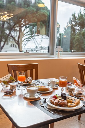 A breakfast table with various food items and drinks, a window with a view of trees, and a chair. - Hotel Villa Maria (Cesenatico)
