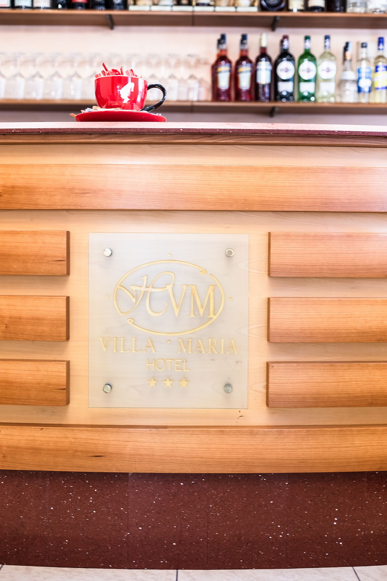 A wooden reception desk with a plaque reading "Villa Maria Hotel" and a row of liquor bottles in the background.