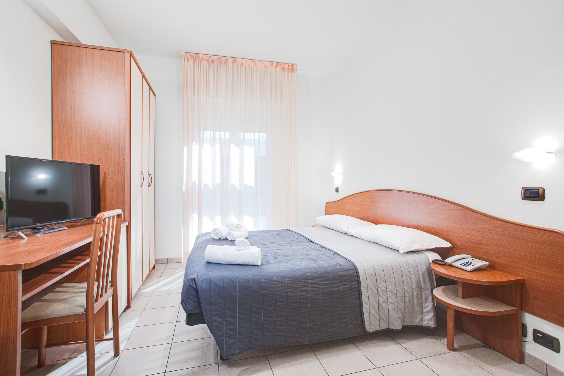 A hotel room with a bed, a desk with a television, a chair, a bedside table with a phone, and a window with curtains.