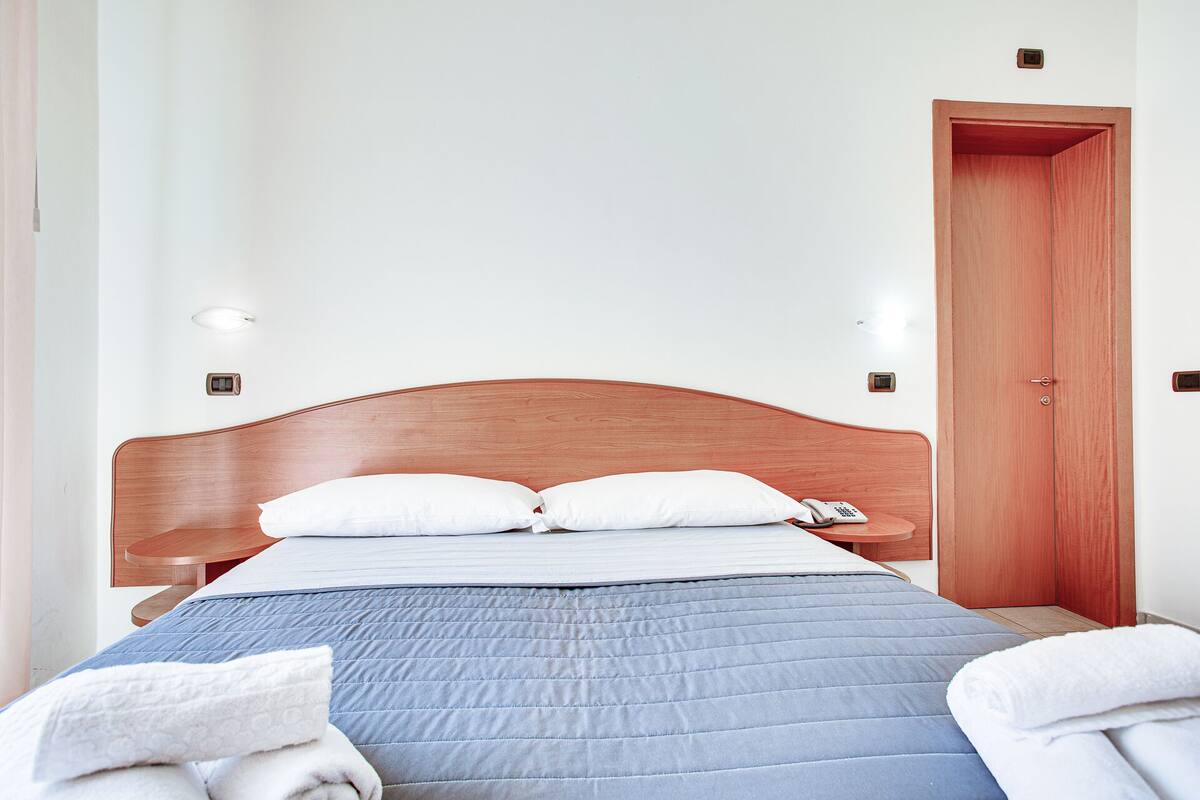 Ein ordentlich bezogenes Bett mit Holzkopfende, weißen Kissen und einer blauen Steudecke. Ein Nachttisch aus Holz mit Telefon und kleiner Lampe steht daneben. Eine Holztür ist rechts sichtbar.