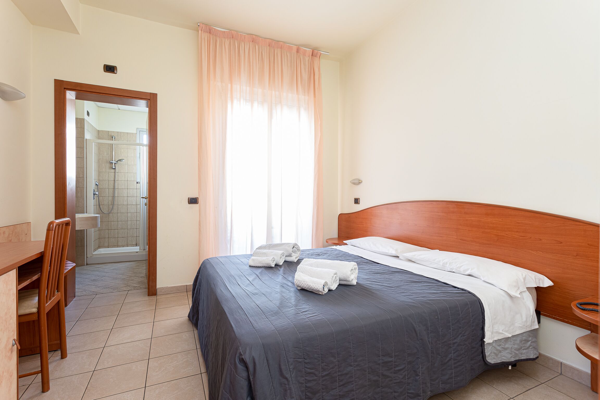 A neatly made bed with a dark blue bedspread and white pillows, a wooden chair, a small wooden table, and a shower area visible through an open door.