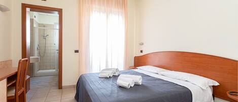 A neatly made bed with a dark blue bedspread and white pillows, a wooden chair, a small wooden table, and a shower area visible through an open door.