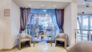 Un área de espera con dos sillones de cuero, una mesa de vidrio con plantas en macetas y vistas a edificios a través de amplias ventanas con cortinas.