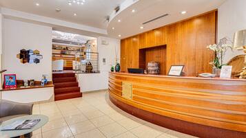 Ein Hotel-Empfangsbereich mit einem Holzbüro, einem Glastisch mit Zeitschriften, einer Treppe, die zu den oberen Stockwerken führt, und einer Bar mit verschiedenen Flaschen.