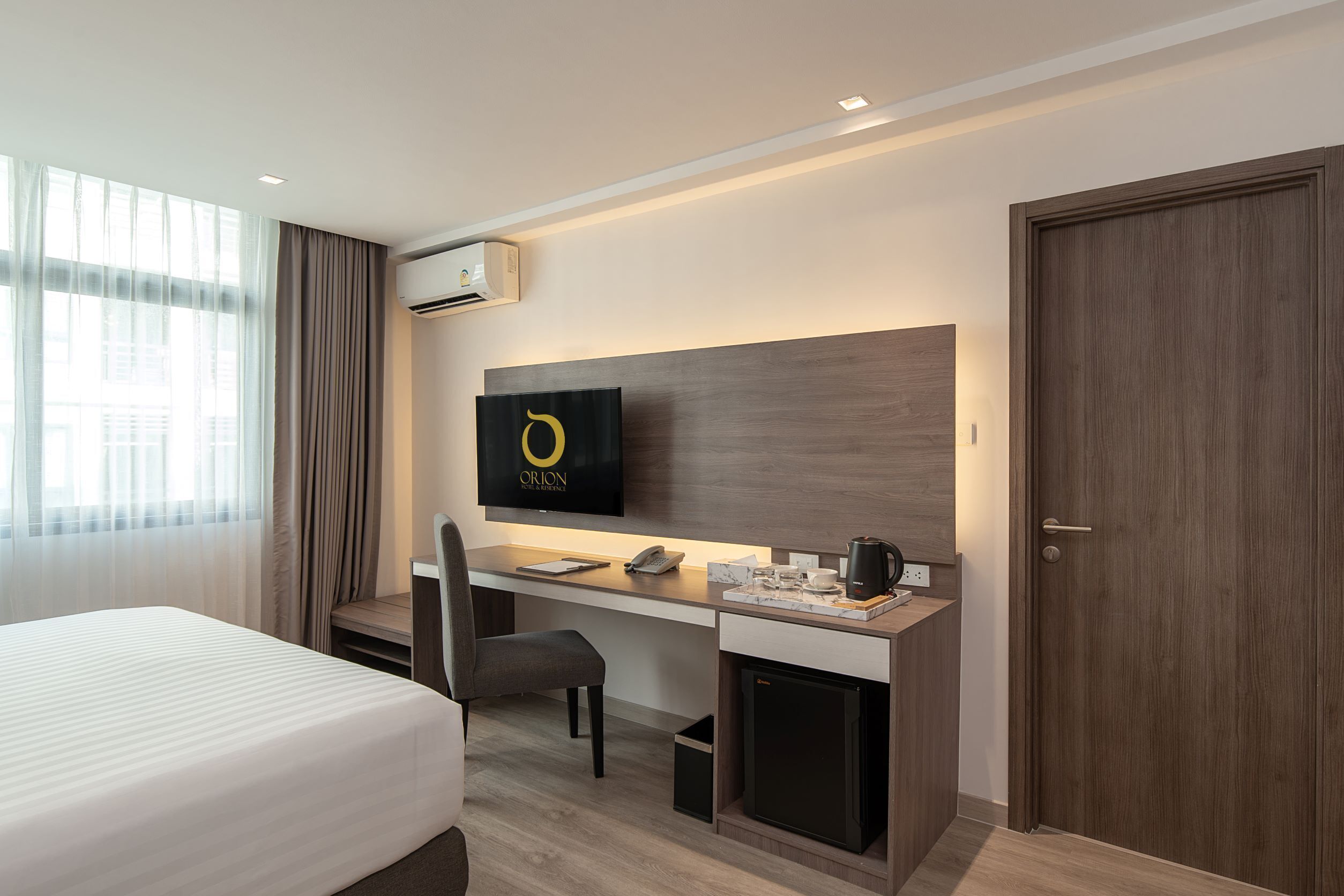 A hotel room with a bed, a desk with a computer, a chair, a small refrigerator, and a TV mounted on the wall.