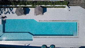 Vista aérea de uma piscina retangular com superfície de água azul cristalina, rodeada por um deck branco com duas guarda-sóis e um espaço de estar.
