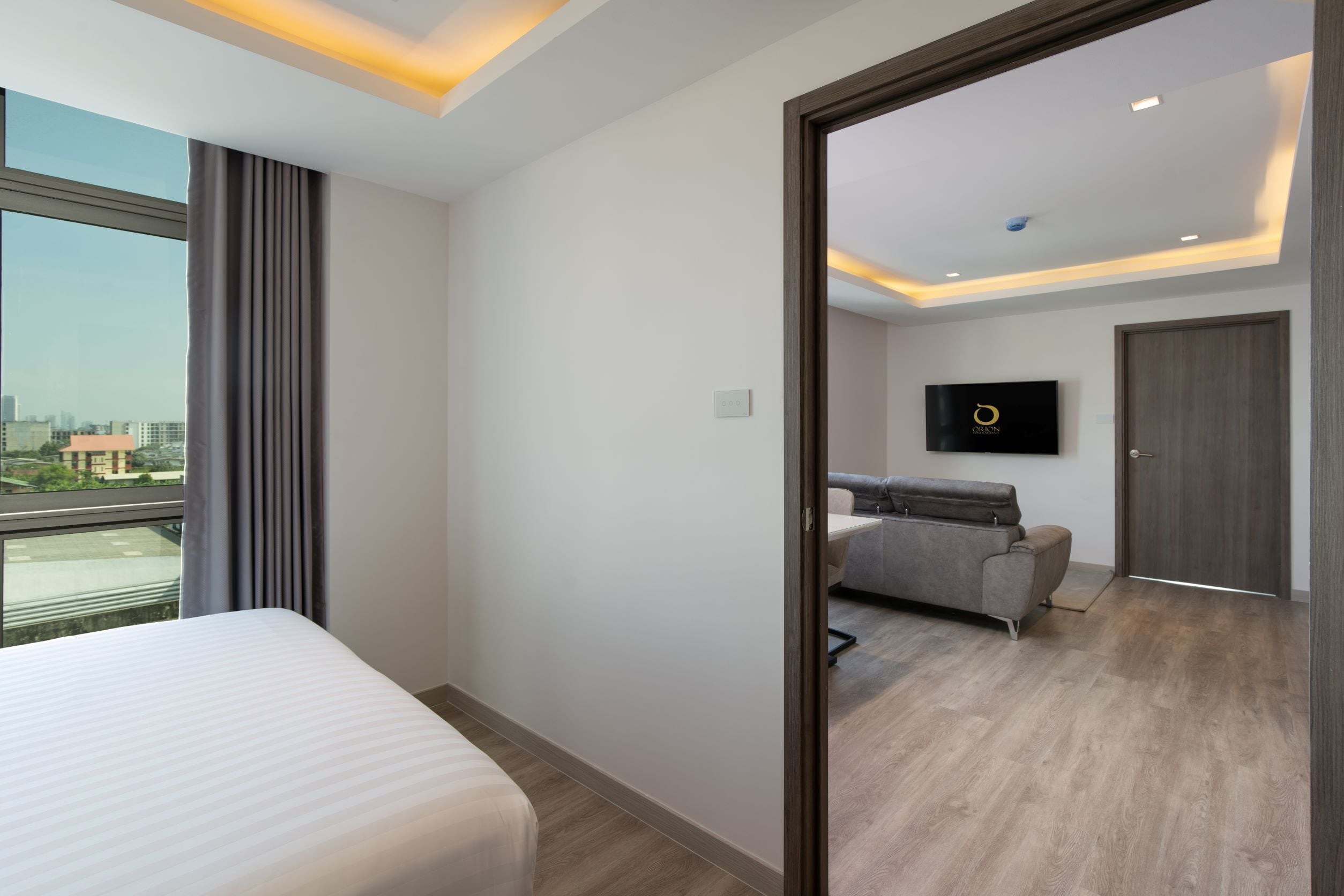 A modern hotel room with a large bed, a sofa, a TV mounted on the wall, and a view of the city through the window.