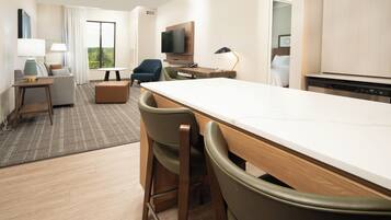 A modern hotel room with a dining area, a sofa, a TV, and a kitchen area.