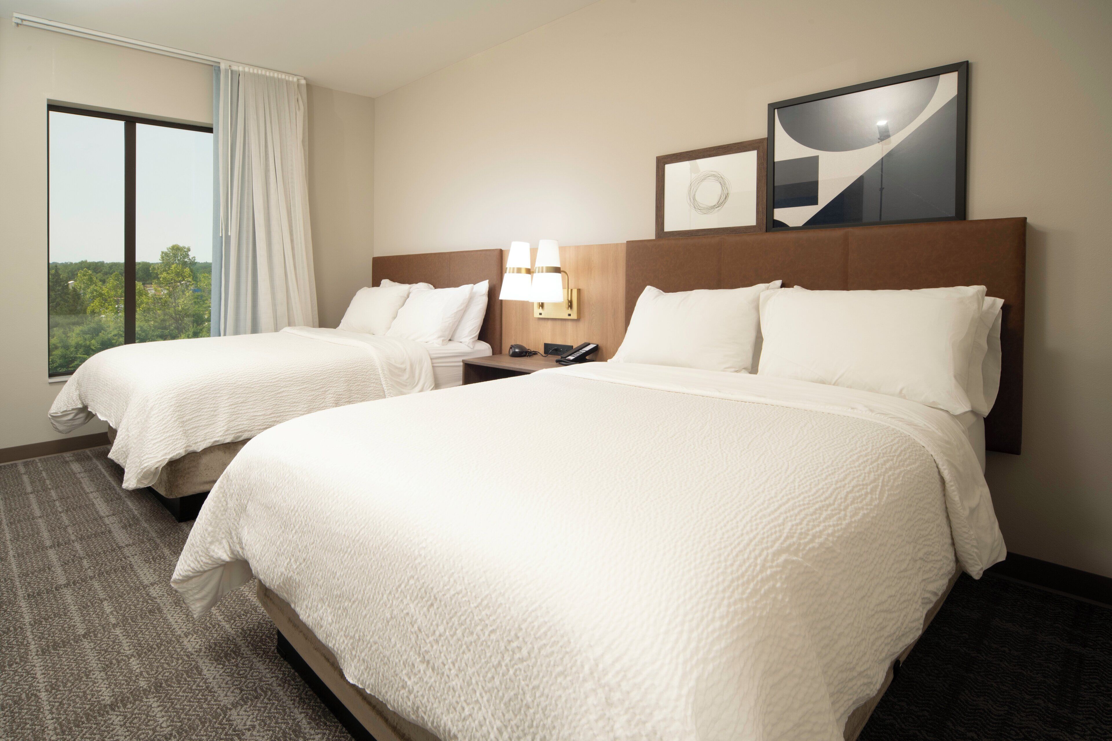 A hotel room with two beds, a large window with a view, a wall-mounted lamp, and a framed picture.