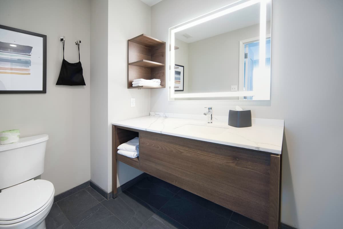 Un baño moderno con un inodoro blanco, un tocador de madera con espejo y una obra de arte enmarcada en la pared.