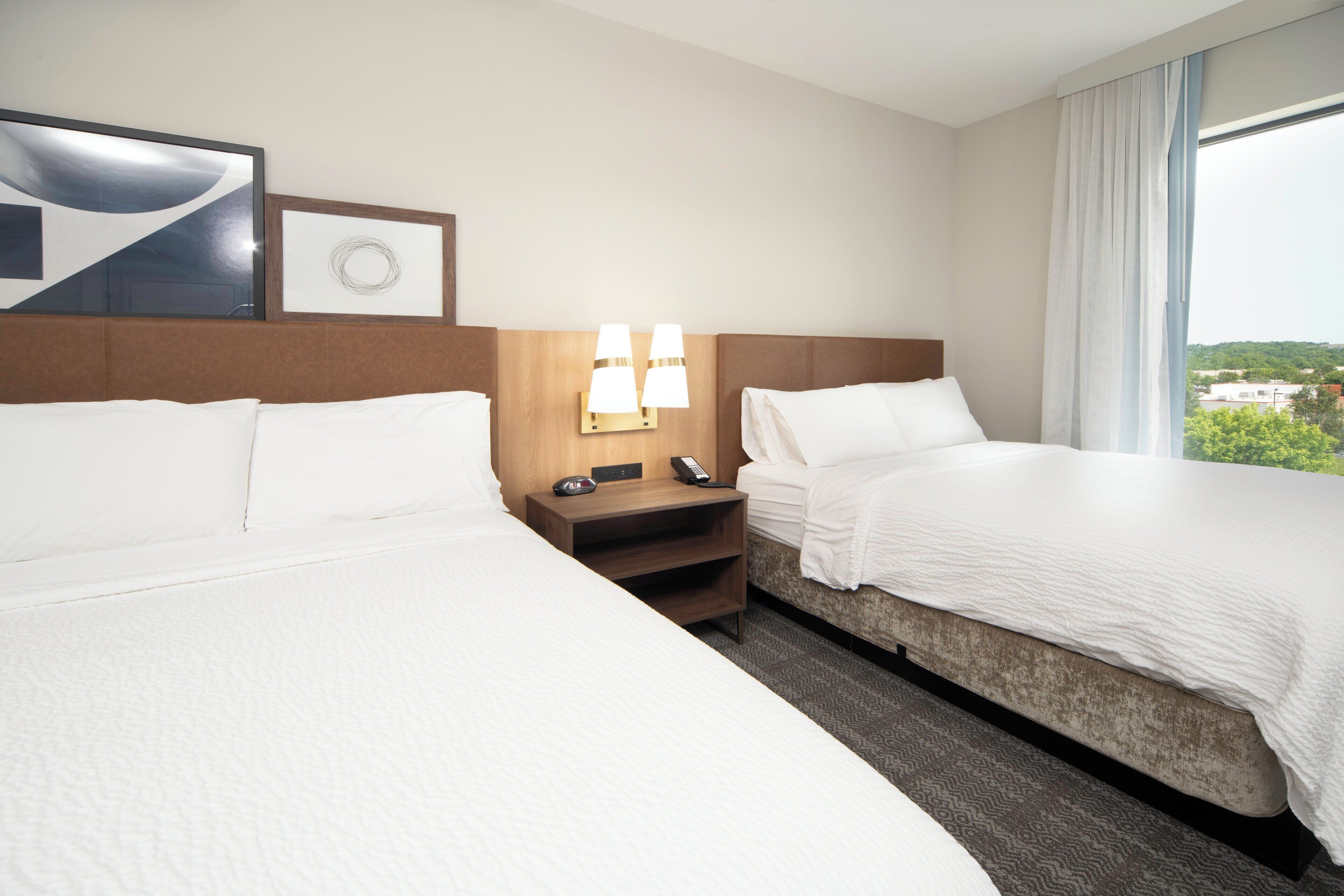 A hotel room with two beds, a nightstand with a lamp, and a view of the city.