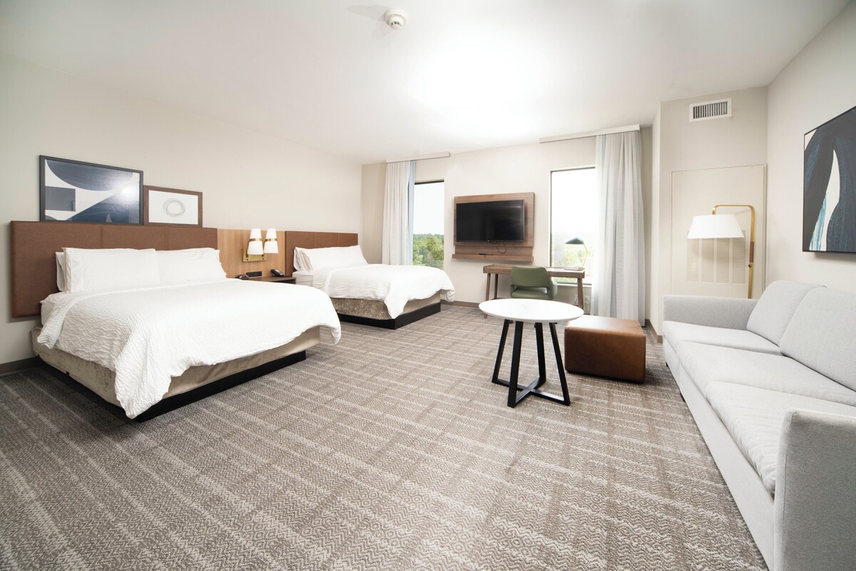 Habitación de hotel con dos camas, un sofá, una mesita, televisor y ventana con cortinas.